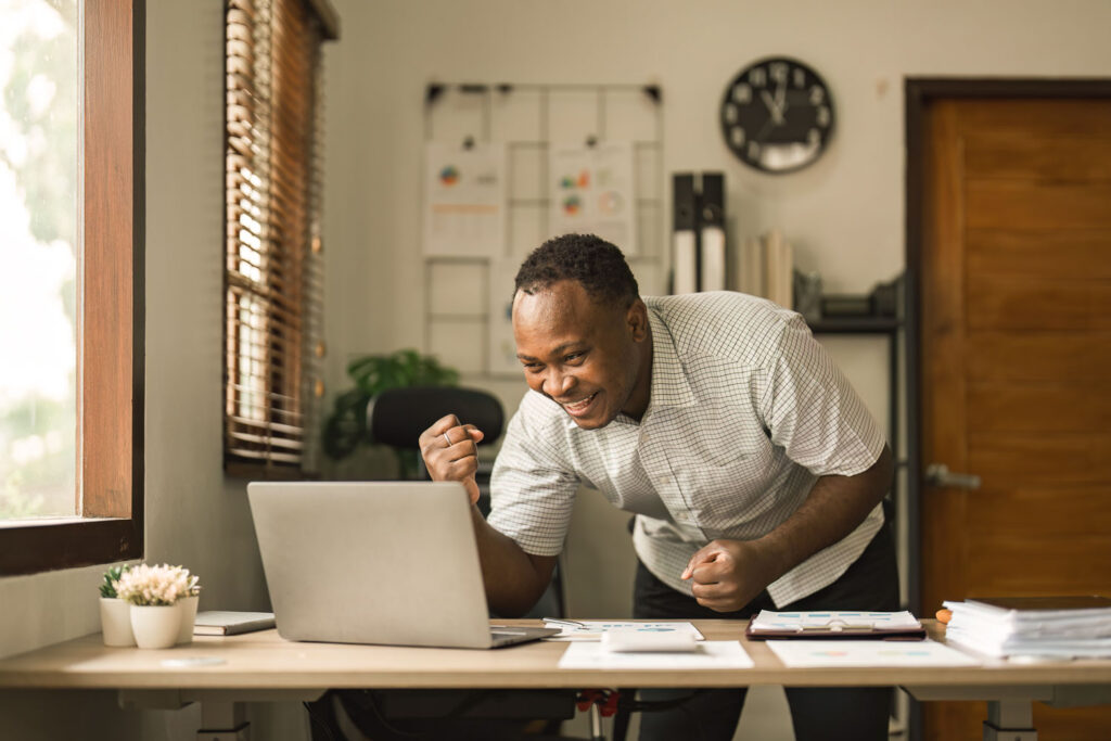 Happy surprised African American businessman reading good news in letter, celebrating success, excited young man holding documents, sitting at work desk
